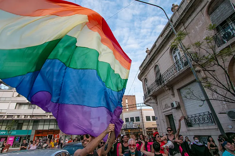 Bandera del arcoíris ondeando en alto durante una marcha del Orgullo en una ciudad de América Latina; activistas celebran en la calle, edificios y cielo parcialmente nublado al fondo.