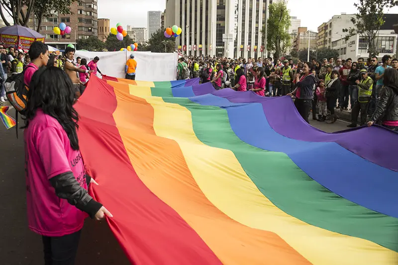 Gran bandera del arcoíris extendida a lo ancho de una avenida; personas la sostienen durante la Marcha del Orgullo en Latinoamérica, con edificios, globos y público alrededor.