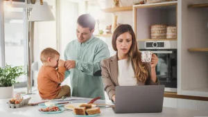 Mujer trabajando con una notebook en una cocina mientras un hombre acompaña a un niño junto a la mesa.