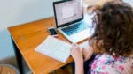 Persona escribiendo en un cuaderno junto a una notebook y un celular sobre una mesa.