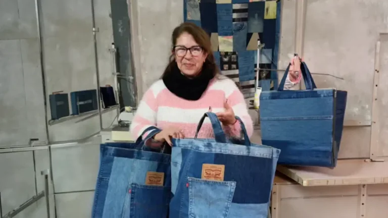 Una mujer sostiene dos bolsos confeccionados con jeans reutilizados en el taller textil de DE LA NADA. Detrás se observan máquinas de coser, retazos de denim y un panel de telas; los bolsos conservan bolsillos y etiquetas de Levi’s.