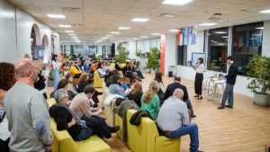 Una expositora habla ante un grupo de personas sentadas en un espacio de trabajo de Natura. Cerca de ella, un hombre opera una computadora; al fondo cuelgan las banderas de Chile, Argentina y Uruguay, entre plantas y sillones verdes.