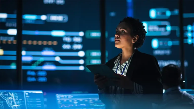 Mujer de piel oscura, con blazer negro y camisa a rayas, sostiene una tablet en una sala de control. A su alrededor, pantallas azules muestran gráficos y datos; la escena vincula tecnología, inteligencia artificial e inclusión laboral.
