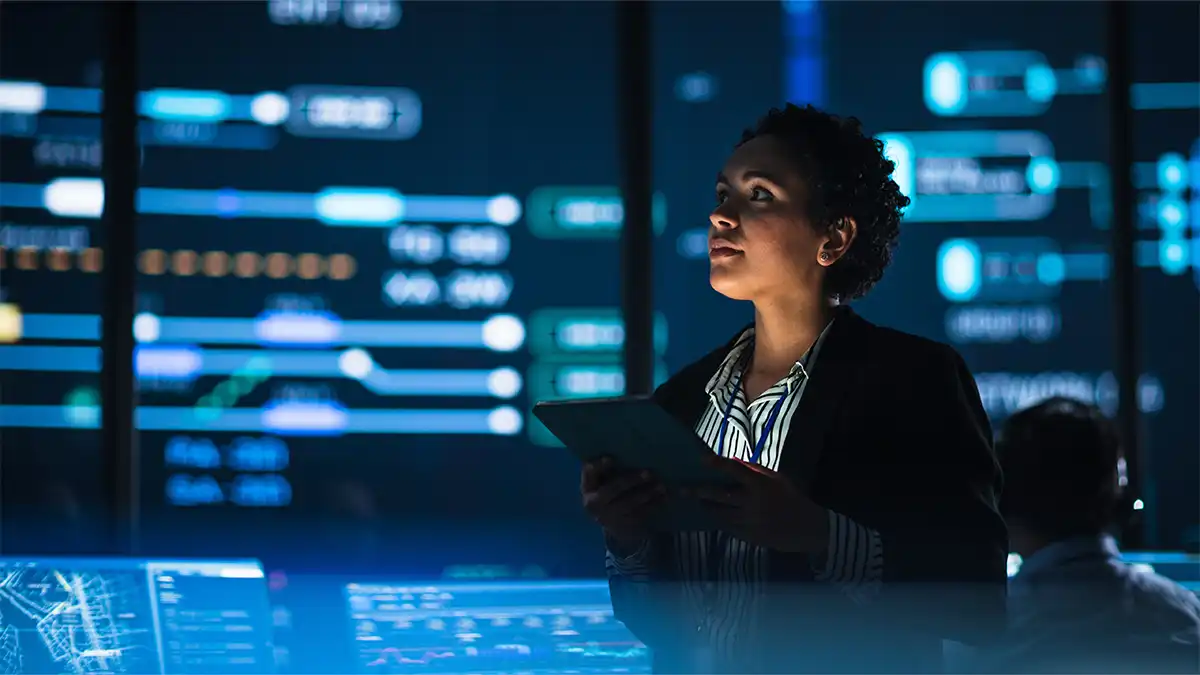 Mujer de piel oscura, con blazer negro y camisa a rayas, sostiene una tablet en una sala de control. A su alrededor, pantallas azules muestran gráficos y datos; la escena vincula tecnología, inteligencia artificial e inclusión laboral.