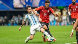 Futbolista de la selección argentina, con la camiseta número 10, disputa la pelota con un rival de camiseta roja número 14 durante un partido. Al fondo aparecen otro jugador y las tribunas desenfocadas; la escena acompaña el debate sobre racismo y Mundial.