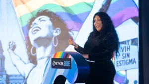 Ana Laura Nicoletti habla en el escenario de GNetwork360 frente a una pantalla con banderas del orgullo y el texto “VIH sin estigma”.