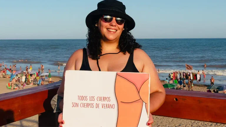 Mujer sonriente con sombrero y anteojos en una playa, sosteniendo un cartel que dice “Todos los cuerpos son cuerpos de verano”.