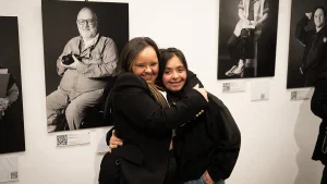 Dos jóvenes se abrazan frente a retratos en blanco y negro de la muestra Entre Retratos en la Alianza Francesa de Buenos Aires.