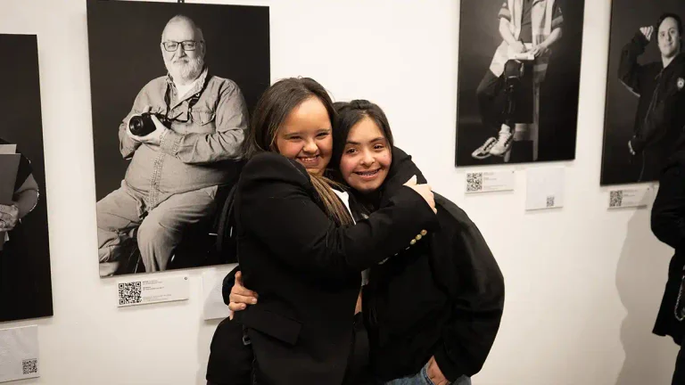 Dos jóvenes se abrazan frente a retratos en blanco y negro de la muestra Entre Retratos en la Alianza Francesa de Buenos Aires.