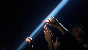 Varias manos y el rostro de una persona se recortan frente a un haz de luz azul en una escena oscura.