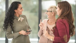 Tres mujeres conversan y sonríen durante un encuentro en un espacio de trabajo; una de ellas sostiene un teléfono celular.