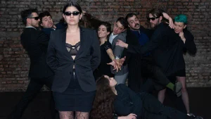 Fotografía del elenco de la obra teatral 1%. En primer plano, una intérprete con traje negro y anteojos de sol mantiene una expresión seria, mientras detrás un grupo de intérpretes vestidos de oscuro adopta gestos y posturas exageradas frente a una pared de ladrillos. Otra intérprete aparece recostada en el suelo.