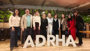Nueve personas posan en un espacio interior frente a grandes letras blancas con la sigla ADRHA. Detrás se observa una pared con vegetación y luces colgantes.