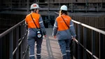 Dos trabajadoras de ArcelorMittal Acindar, con cascos blancos y uniformes grises y naranjas, caminan por una pasarela de una planta industrial.