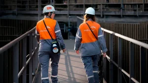 Dos trabajadoras de ArcelorMittal Acindar, con cascos blancos y uniformes grises y naranjas, caminan por una pasarela de una planta industrial.