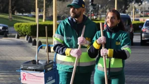 Dos trabajadores de Cliba posan con escobas junto a un carro de limpieza con cartelería de Octubre Rosa, en una acción de concientización sobre el cáncer de mama.