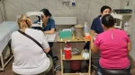 Dos mujeres reciben atención oftalmológica en un consultorio. Frente a ellas, profesionales de salud utilizan equipos de examen ocular. Entre ambos puestos hay una mesa con instrumental médico.