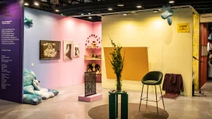 Espacio expositivo de ELA en arteba 2025, con paredes violetas, rosadas y amarillas, pinturas, objetos artísticos, esculturas textiles, una silla verde y un arreglo vegetal sobre una mesa.