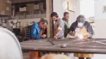En un taller de metalurgia, una persona con máscara y delantal protector realiza una soldadura sobre una estructura metálica, mientras otras personas observan el trabajo y toman notas.