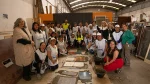 Grupo de mujeres participantes y representantes de la capacitación ConstruACTORAS posan en un taller de materiales de construcción, junto a superficies de pisos y revestimientos trabajadas durante la jornada práctica.