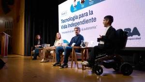 Cuatro panelistas participan de una conversación sobre tecnología para la vida independiente. En primer plano, una persona interviene desde una silla de ruedas motorizada. Detrás se proyecta una pantalla con el título «Tecnología para la Vida Independiente».