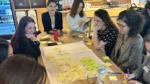 Mujeres profesionales participan de un taller colaborativo de Juntas+, reunidas alrededor de una mesa con notas adhesivas amarillas y verdes sobre una hoja de trabajo, cuadernos y materiales.