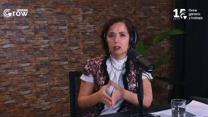 Una mujer con auriculares y chaleco estampado habla ante un micrófono durante una transmisión de los Premios Grow 2026. Detrás se observa una pared revestida en piedra y, en las esquinas superiores, los logotipos de Premios Grow y los 15 años de Grow - Género y trabajo.