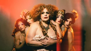 Cinco intérpretes de Saraos Uranistas posan con pelucas voluminosas, maquillaje teatral y vestuario de estética cabaretera. En el centro, un personaje de cabello rojizo rizado y vestido claro mira a cámara, rodeado por cuatro personajes bajo una iluminación anaranjada.