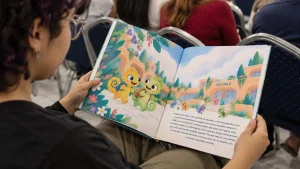 Persona con anteojos sostiene abierto un ejemplar del cuento infantil Tito, el camaleón amarillo. En las páginas se observan dos camaleones ilustrados, uno amarillo y otro verde, rodeados de plantas y flores, frente a una escuela. Al fondo hay otras personas sentadas.